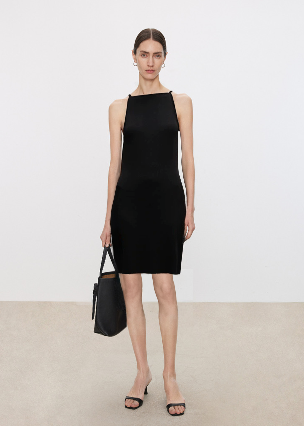 Woman wearing a black sleeveless dress holding a black handbag against a plain background