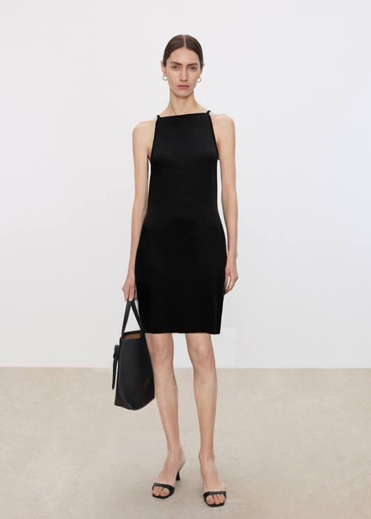 Woman wearing a black sleeveless dress holding a black handbag against a plain background